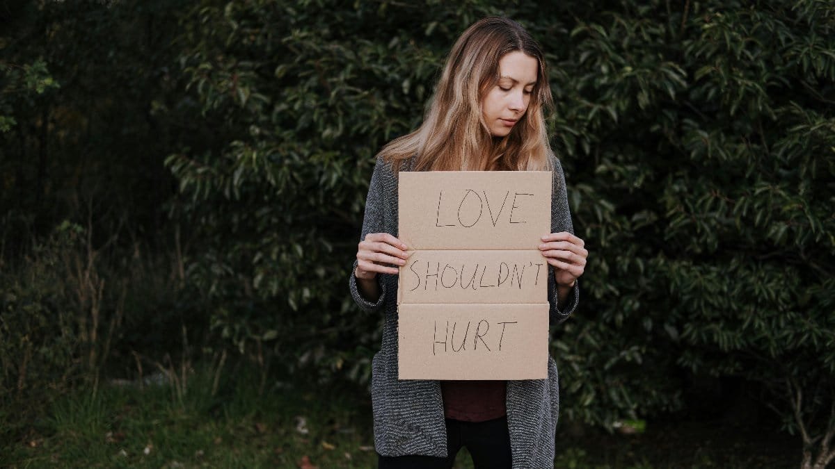 Lonely upset female in casual outfit standing near trees and grass while holding cardboard against abuse with inscription Love Should Not Hurt