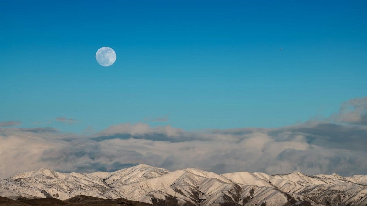 Capture of snow-covered mountains under a blue sky with the moon in view, showcasing nature's serene beauty.