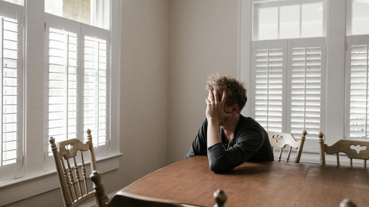 A man sits alone at a table in a bright room, displaying deep contemplation.