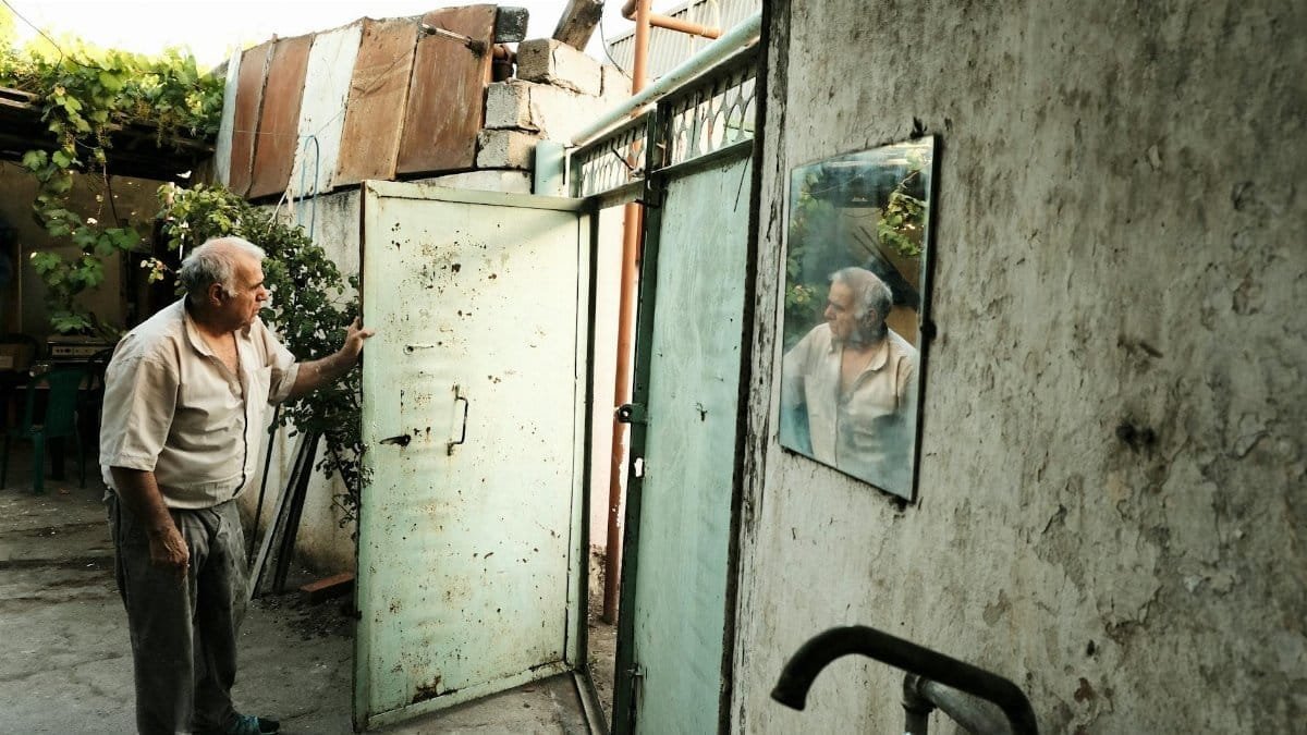 An elderly man opens a worn door in an urban courtyard, reflecting in a mirror on the wall.