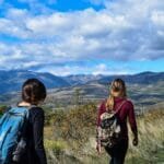 Two women hiking on a mountain trail with scenic views under a blue sky.