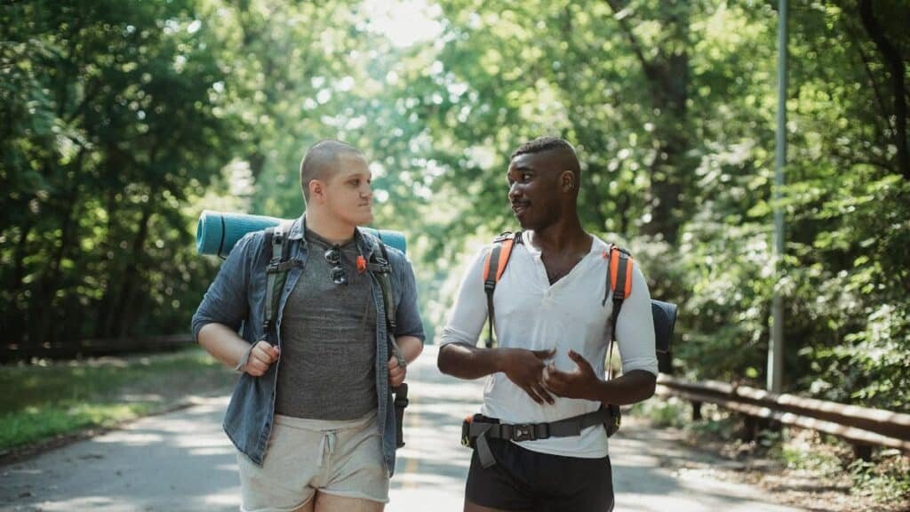 Two friends hiking and talking on a sunny day through a forest road.