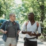 Two friends hiking and talking on a sunny day through a forest road.