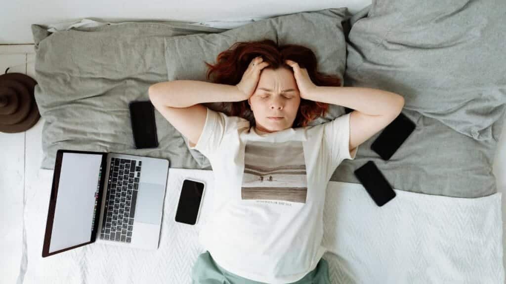A distressed woman lying in bed surrounded by digital devices, showing signs of stress.