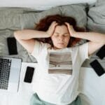 A distressed woman lying in bed surrounded by digital devices, showing signs of stress.