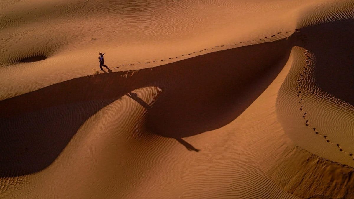 A lone person walks across sand dunes at sunset, casting long shadows in the desert.