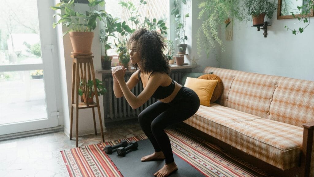 Woman in active wear doing squats on a yoga mat in a cozy indoor setting with plants and natural light.
