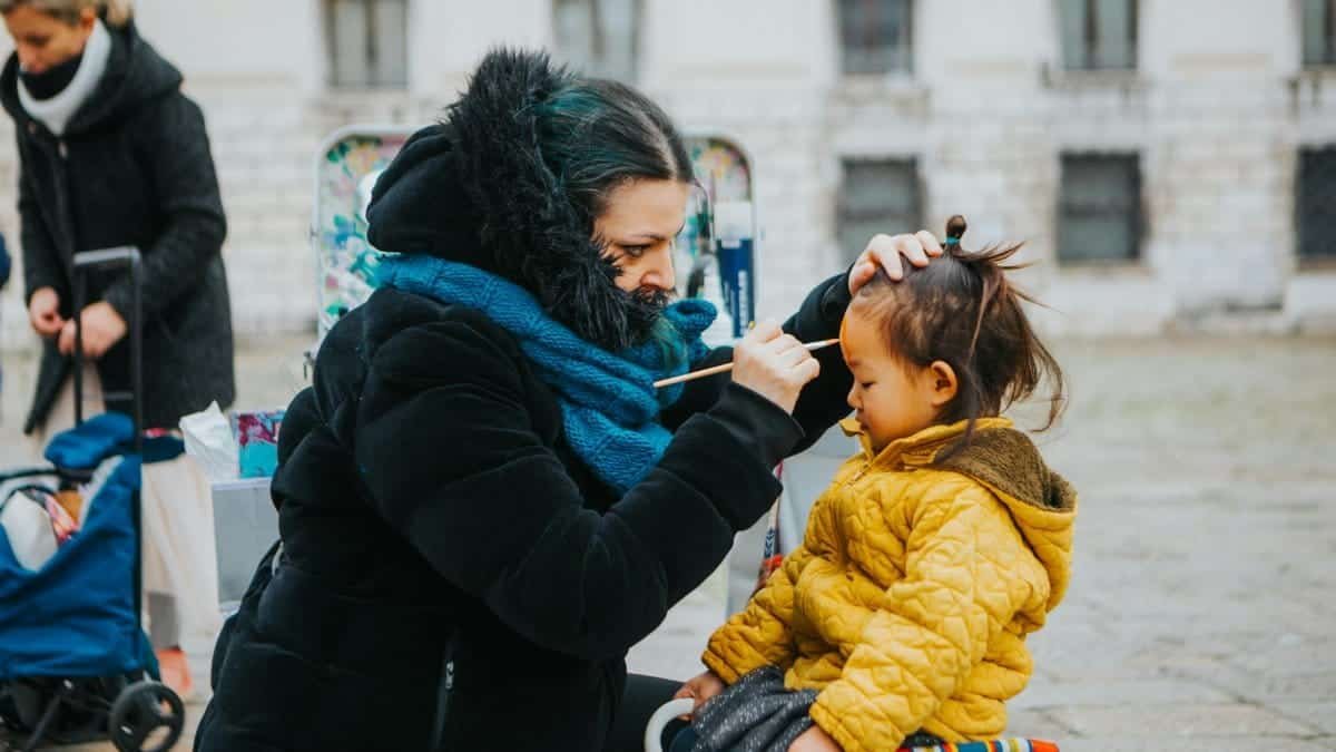 A woman paints a child's face outdoors during a chilly winter day.