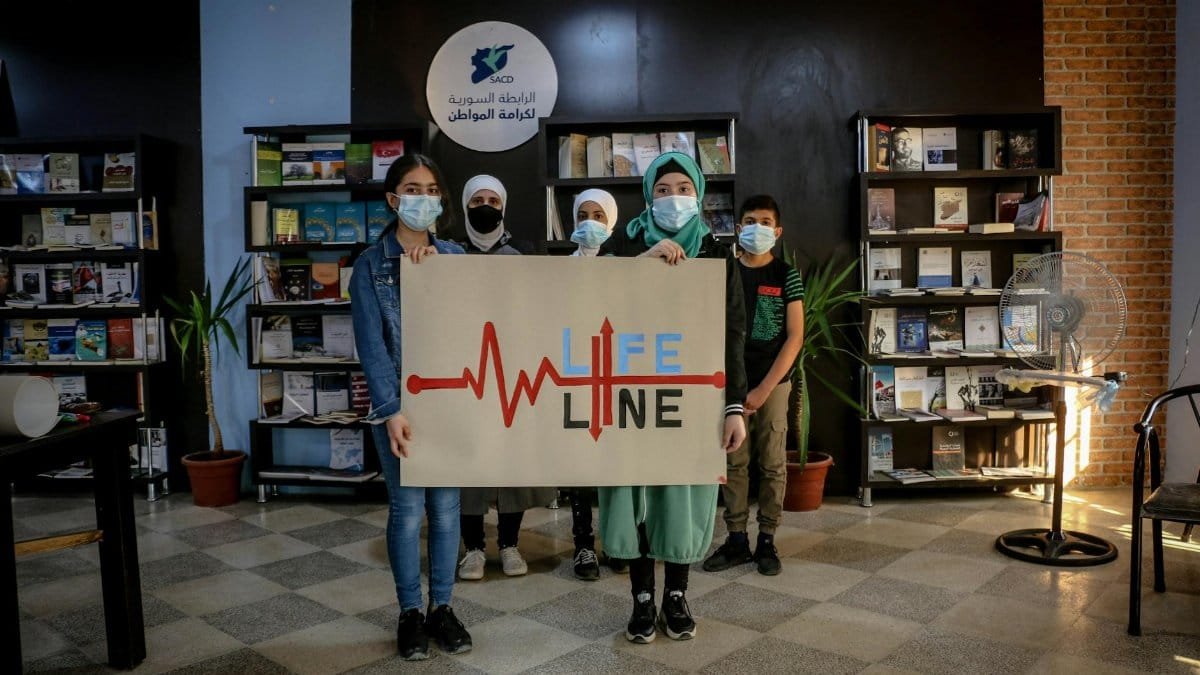 Group of young people holding Lifeline sign in a Syrian library setting.