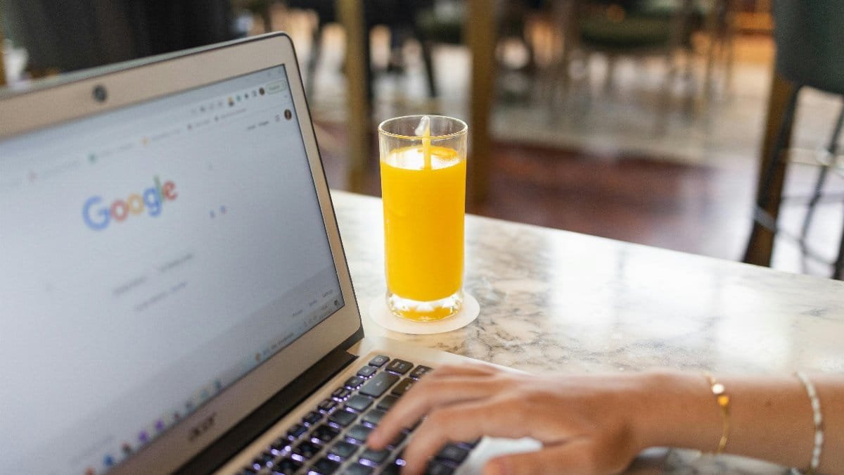Woman typing on laptop with Google homepage open, beside a glass of orange juice on a marble table.