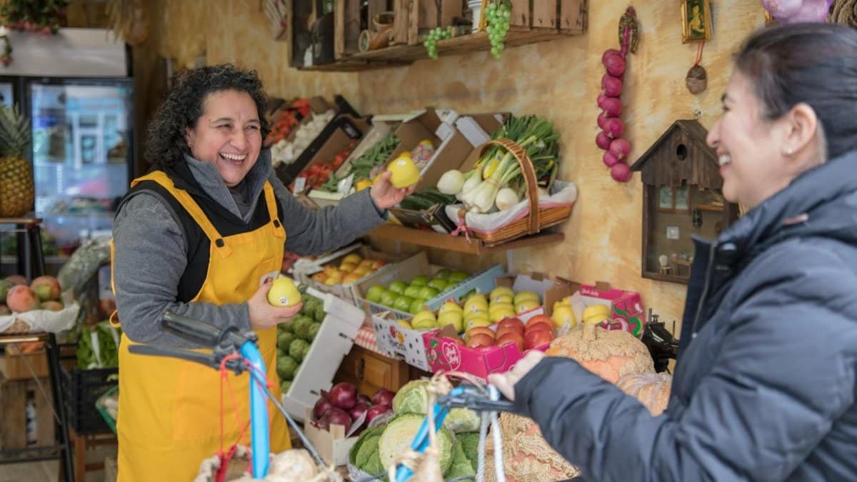 Two women smiling and exchanging fresh produce at a vibrant greengrocer's market.