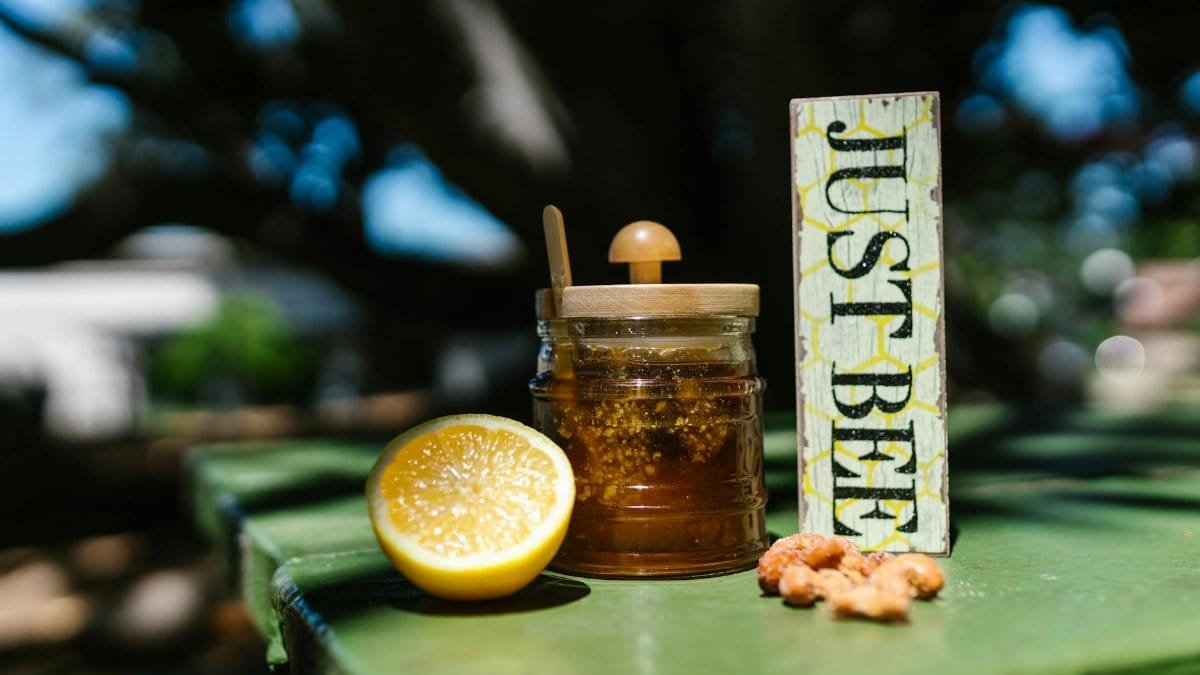Close-up of a honey jar, lemon, and 'Just Bee' sign on a green table outdoors.