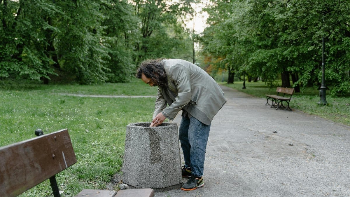 A man searching a trash bin in a city park, highlighting urban poverty.