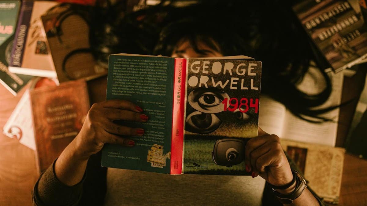 A person lying down, holding and reading George Orwell's 1984, surrounded by scattered books.