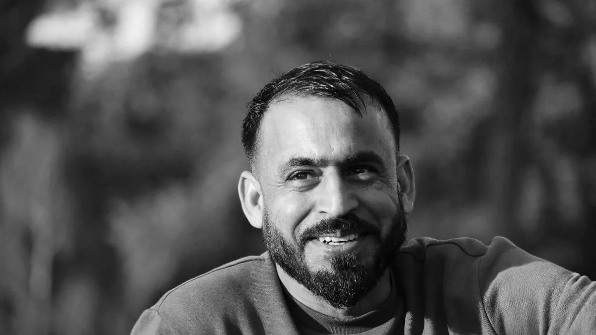 A candid black and white portrait of a smiling man with a beard, set against a blurred outdoor background.