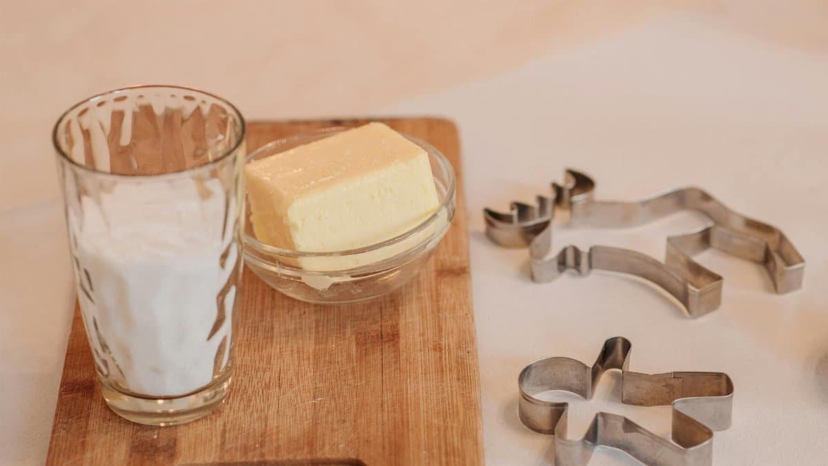 A glass of milk and butter bowl placed on a wooden board with cookie cutters, ready for baking.