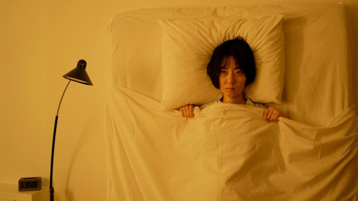 Woman lying under a blanket with a moody expression in a dimly-lit bedroom.