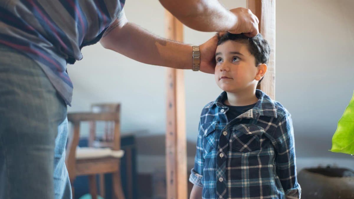 A father marking his young son's height on a wooden pole indoors, capturing a family bonding moment.