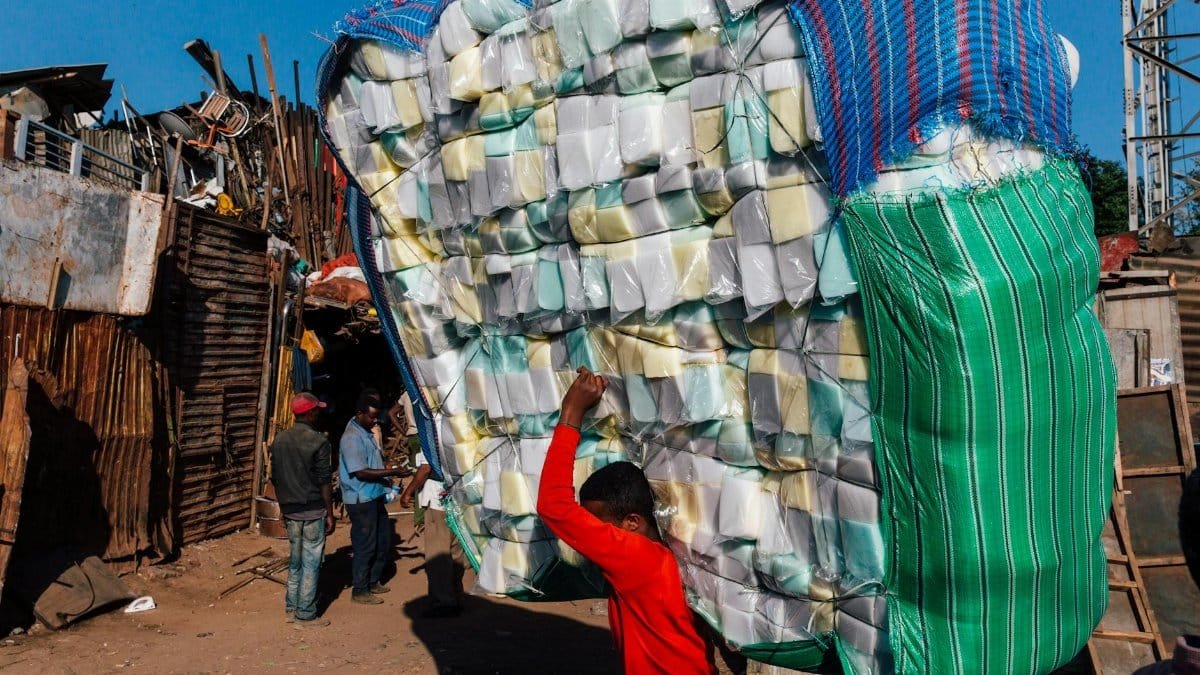 Side view of anonymous ethnic male in casual wear carrying heavy stack of merchandise with striped bags on dry terrain under blue sky in sunlight