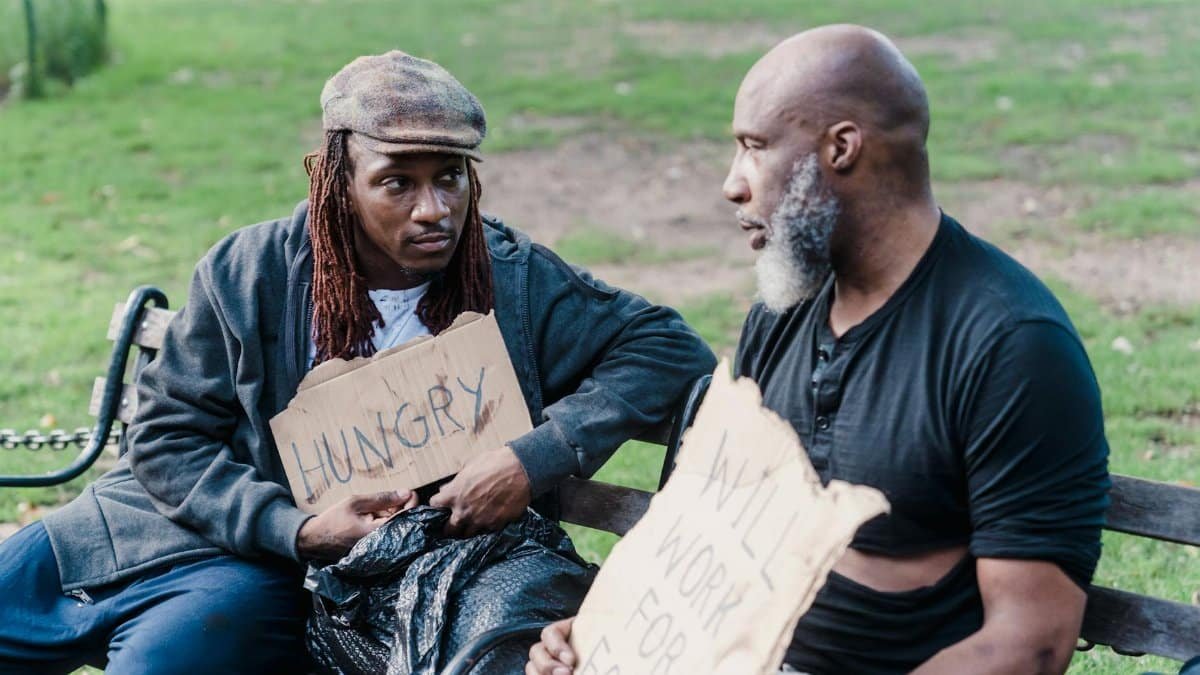 Two men sitting on a bench in a park holding signs about hunger and work.
