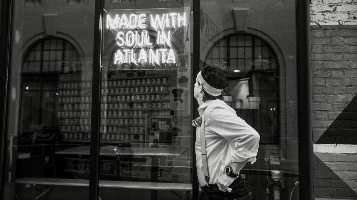 A man in suspenders looks at a neon sign reading 'Made with Soul in Atlanta.' Black and white street photo.