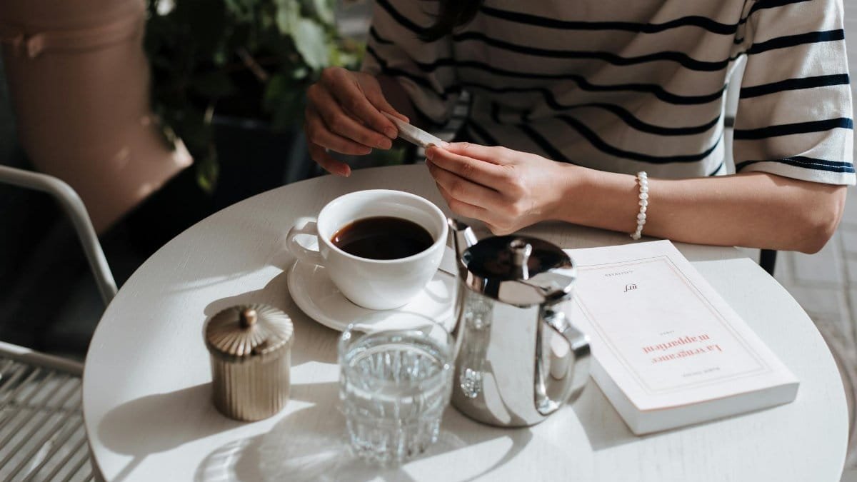 A person enjoying a coffee break, holding a sugar packet next to a cup of coffee and book on a sunny table.