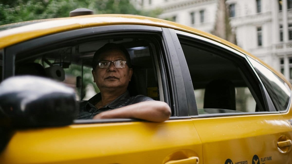 Through window of male driver in glasses sitting in cab while driving in downtown of city and looking away