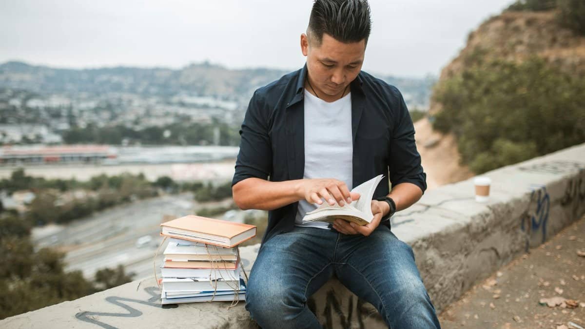 An adult man reads a book outdoors on a concrete wall with a stack of books beside him.