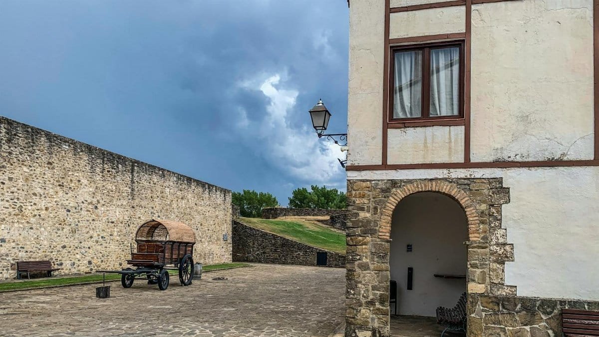 A rustic courtyard scene featuring a historic wagon and stone architecture under dramatic stormy skies.