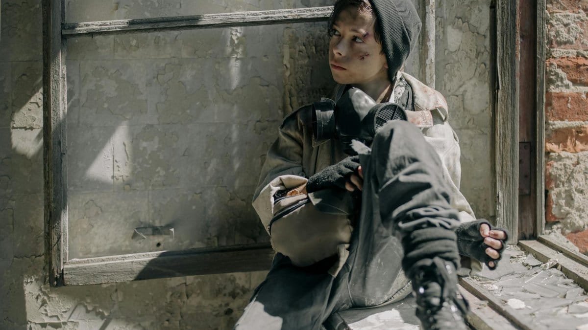 A young boy, dressed in survival gear, sits in a ruined building reflecting desolation and survival.