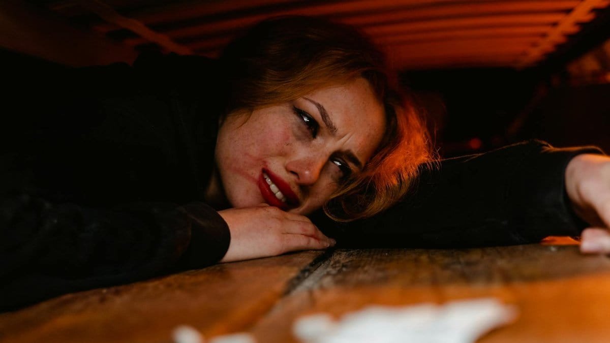 A distressed woman lying under a wooden structure, expressing fear and anxiety.