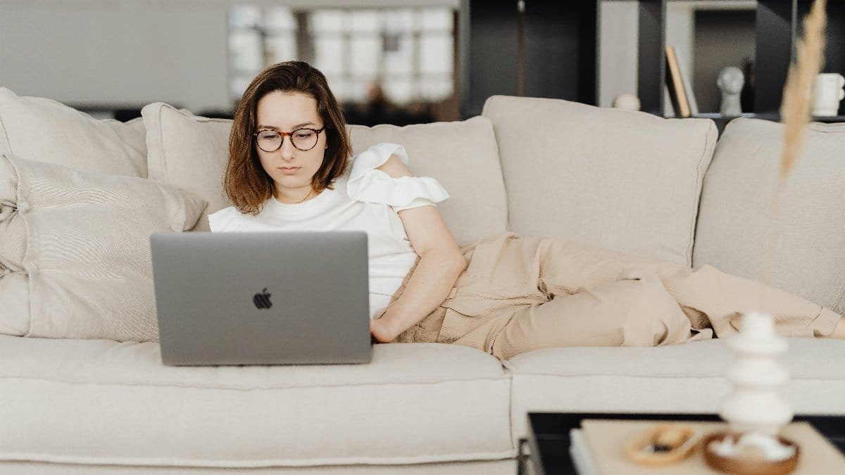 Woman wearing glasses relaxing on a beige couch while using a laptop in a modern living room.