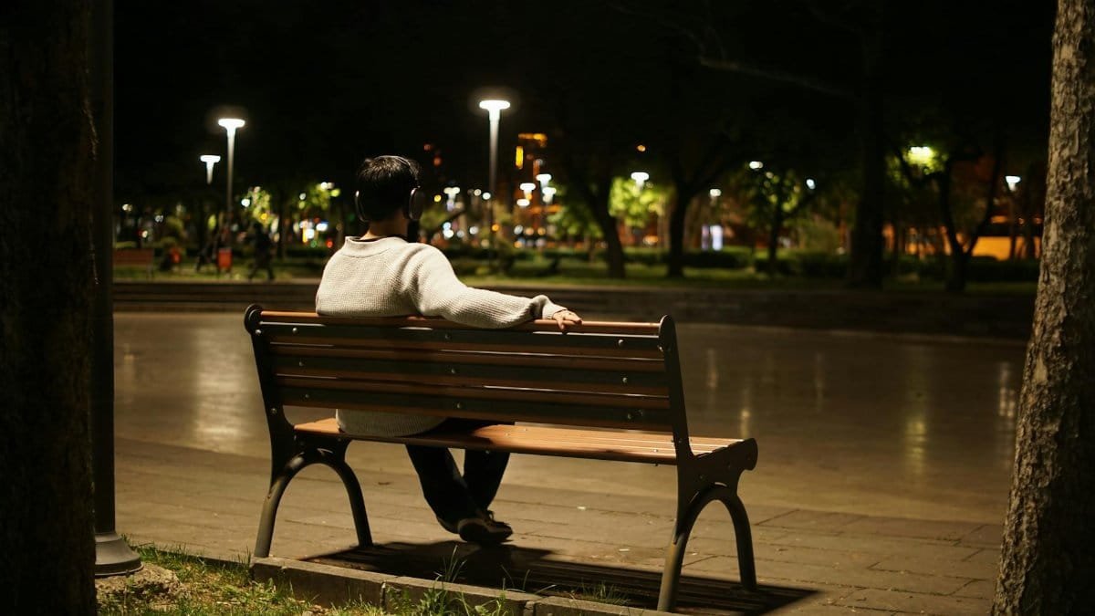 Solitary figure listening to music on a park bench under night streetlights, urban solitude.
