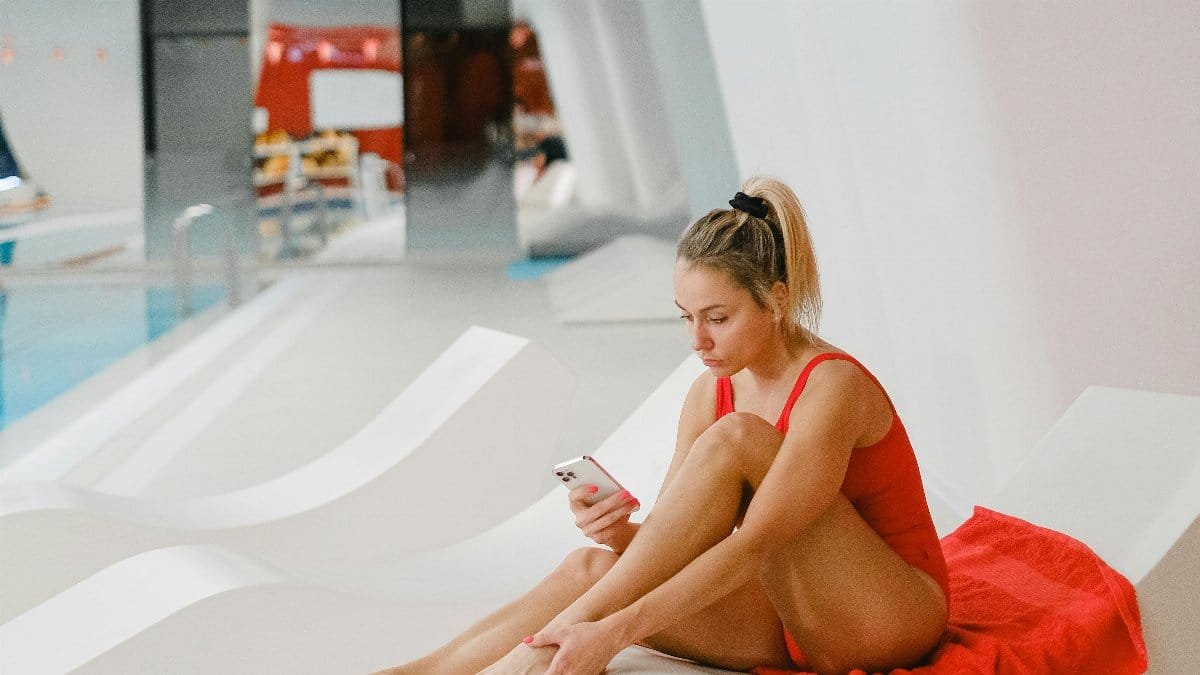 Woman in red swimsuit using smartphone on white poolside lounger indoors.