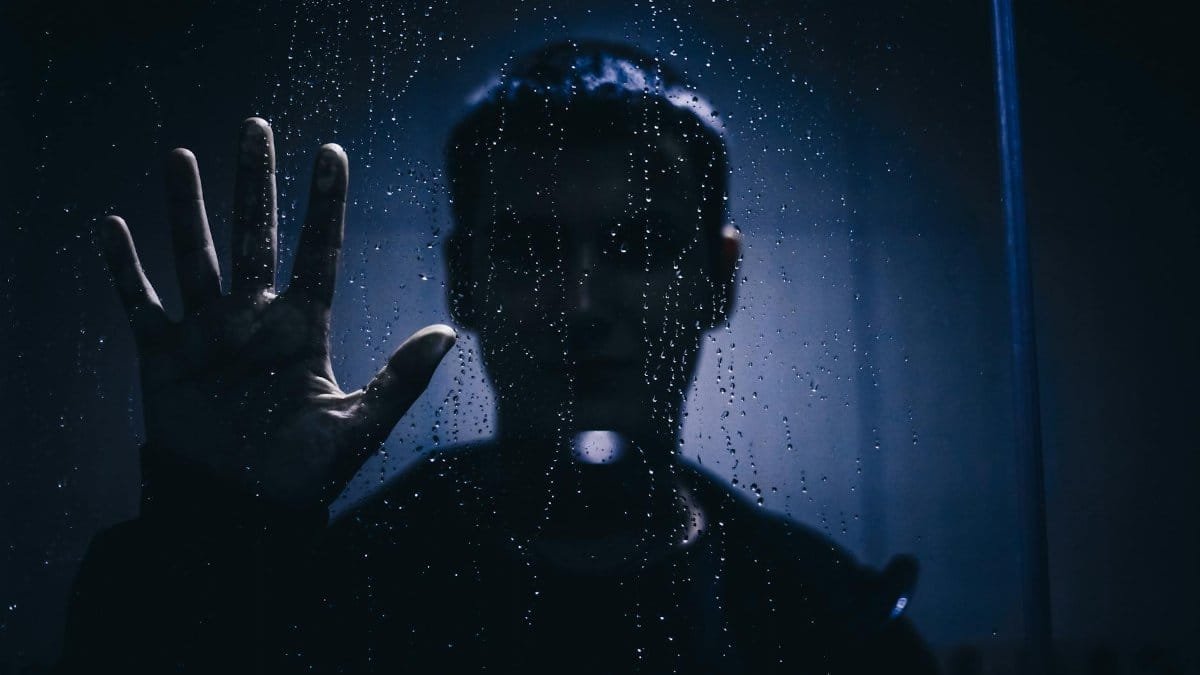 Silhouette of a person with hand against a rain-covered glass, evoking mystery and introspection.