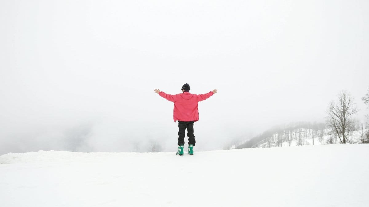 A person in a pink jacket stands with arms wide open on a snowy mountain, embracing the winter scenery.