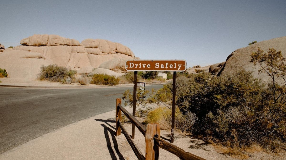 A scenic desert road with a 'Drive Safely' sign surrounded by rocky formations and dry vegetation.