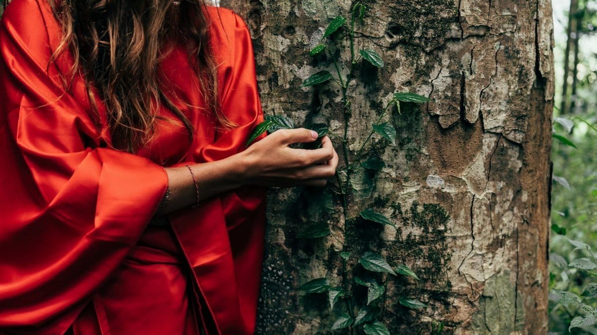 A woman in a vibrant red dress interacts with a lush forest tree, evoking connection with nature.