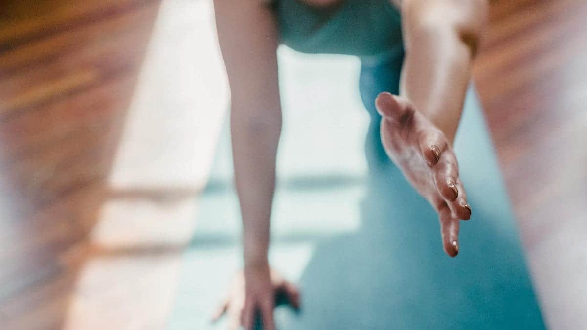 Person practicing yoga on a mat indoors, reaching forward in natural sunlight.