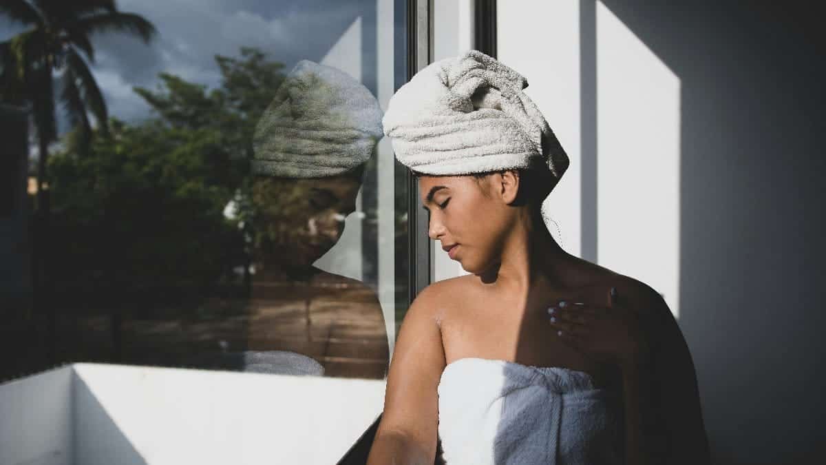 A woman with a towel by a glass window, her reflection visible, creating a serene and contemplative mood.