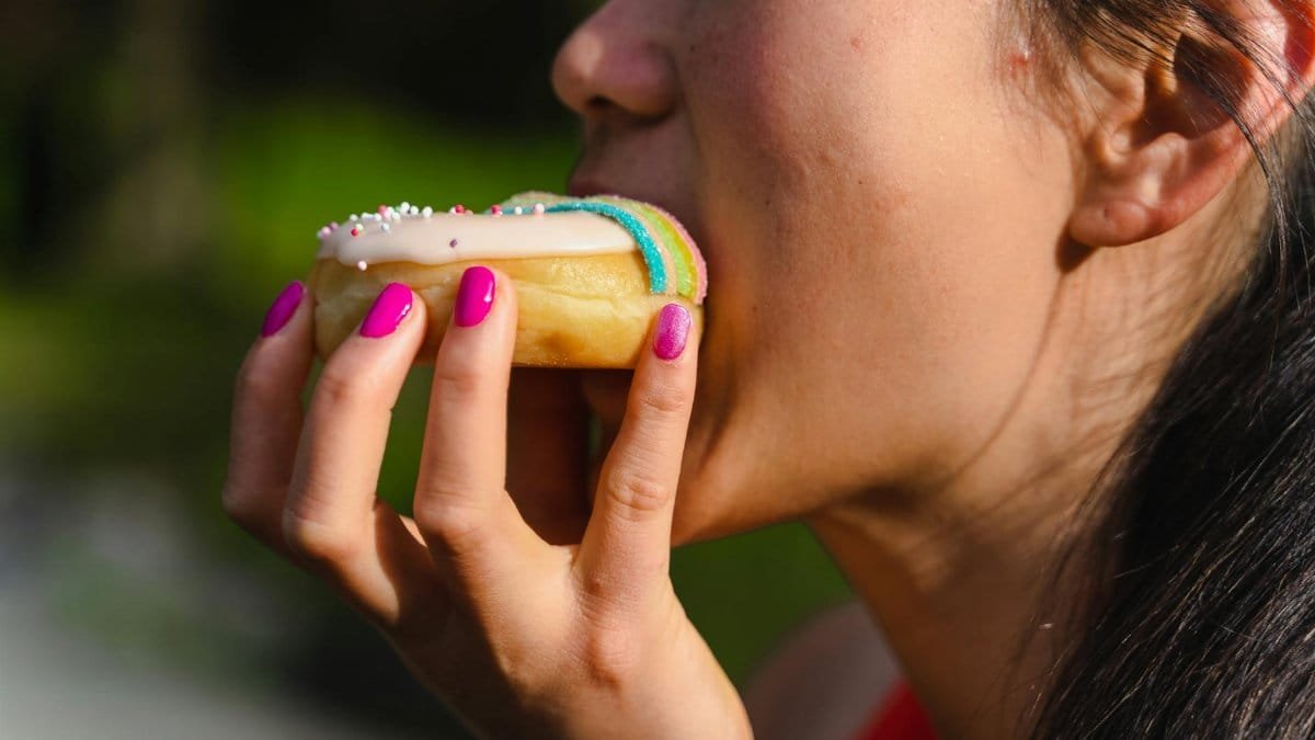 A woman with manicured nails takes a bite of a colorful sprinkled doughnut.