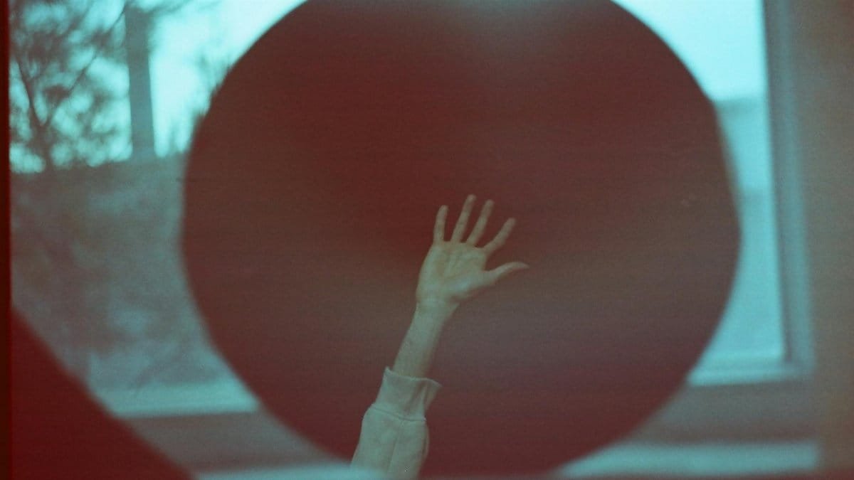 An artistic image featuring a hand raised against a circular reflective surface, captured with film photography.