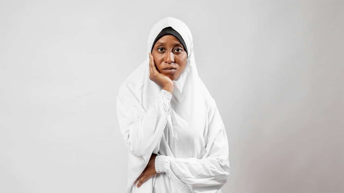 A portrait of a thoughtful woman wearing a traditional white hijab against a plain background.
