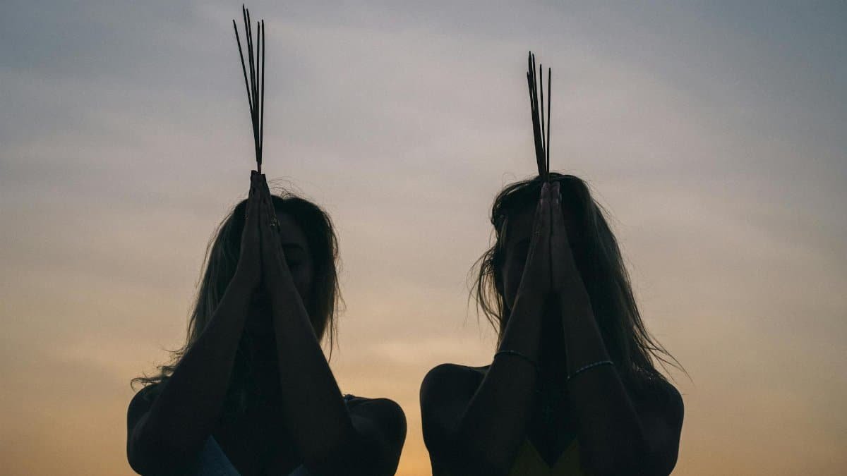Two women meditating at sunset with incense sticks, creating a calming silhouette scene.