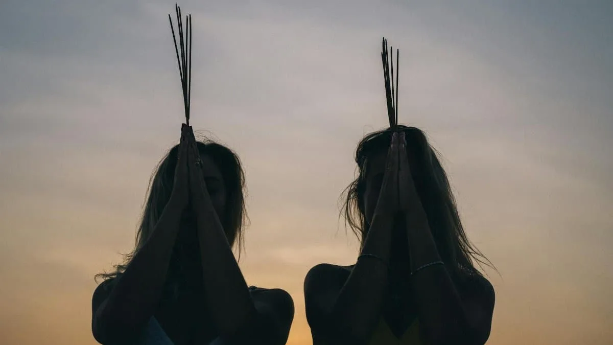 Two women meditating at sunset with incense sticks, creating a calming silhouette scene.
