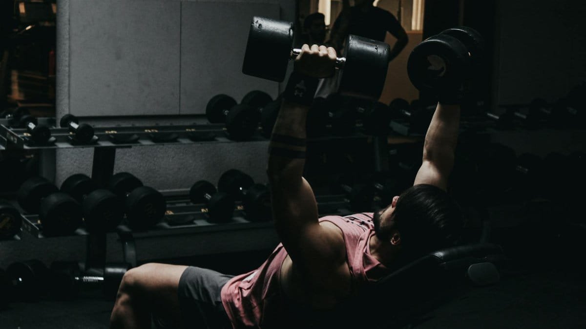 Muscular man in sportswear performing dumbbell bench press in a dimly lit gym setting.
