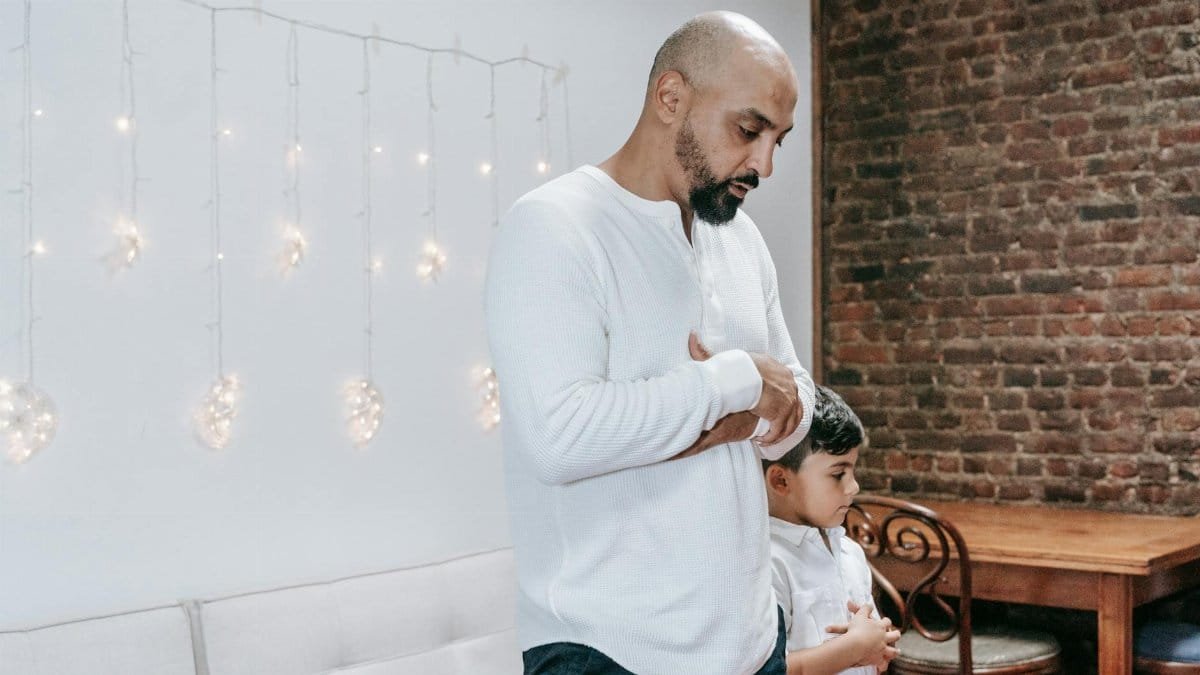 Father and child in thoughtful prayer indoors, beneath gentle string lights.