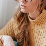 Pensive young female student in warm outfit and eyeglasses sitting with book while looking away in light white room via Pexels
