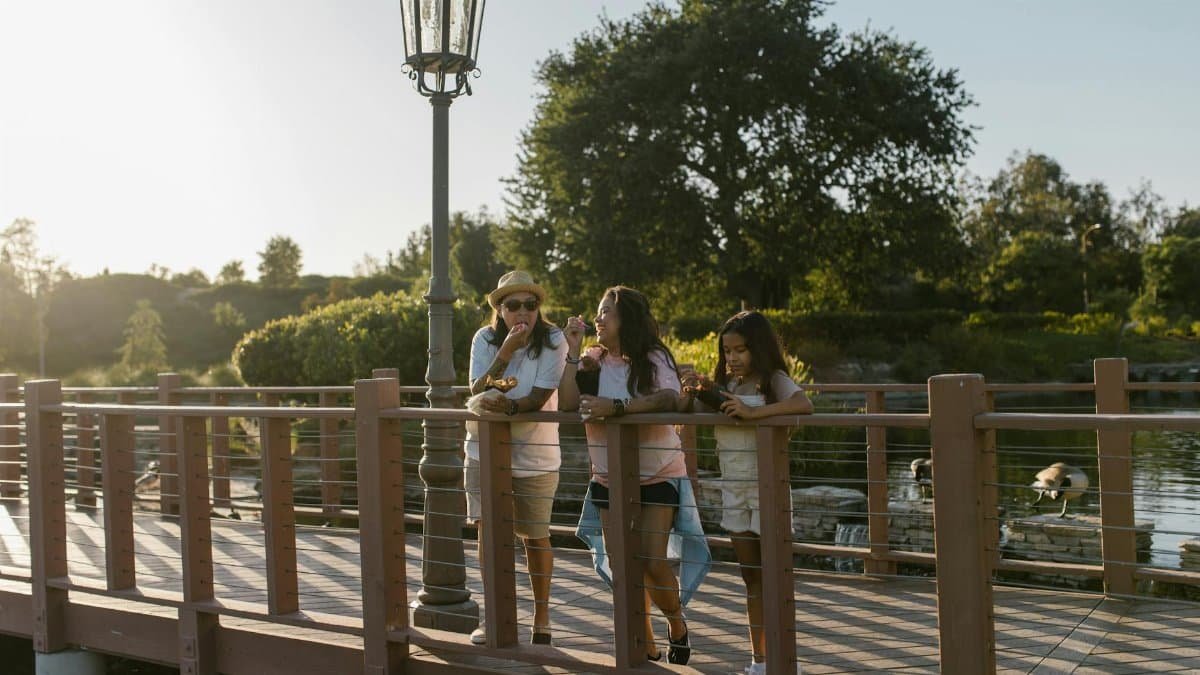 A family enjoys snacks on a sunny footbridge with scenic nature in the background.