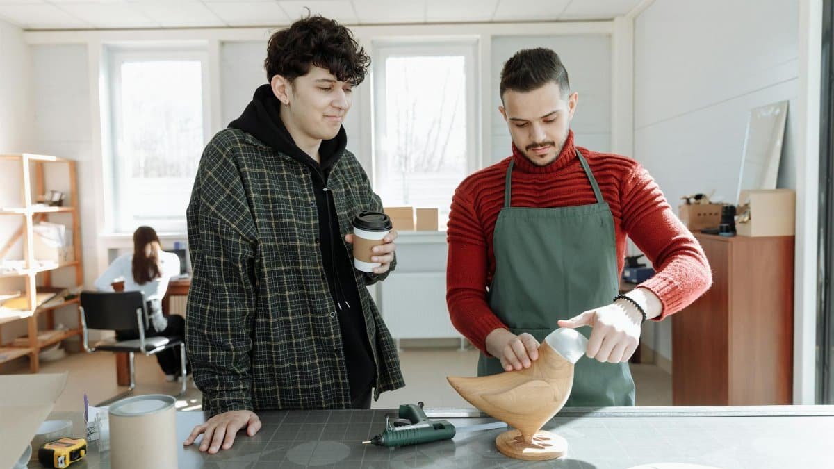 Two young men crafting a wooden figurine in a sunlit workshop. Teamwork and artistry indoors.